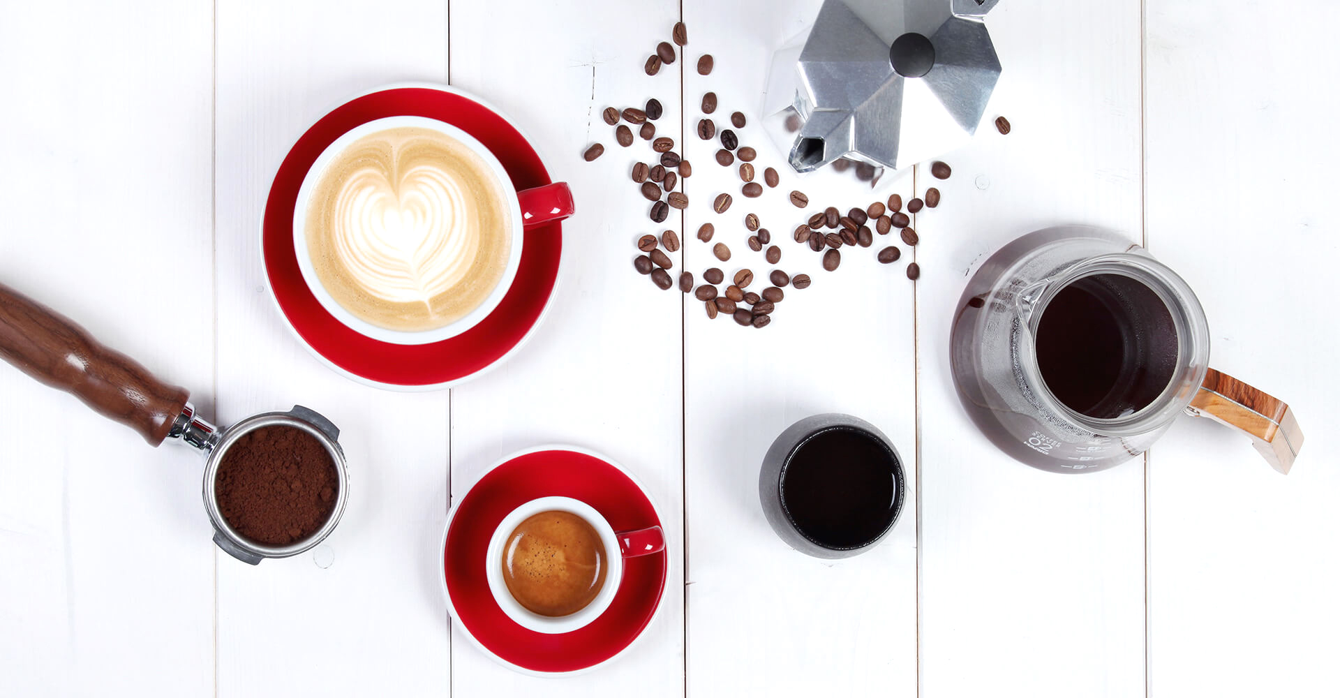 Eine rote Cappuccinotasse mit Latte Art neben einer roten Espressotasse, dazu ein gefüllter Siebträger, eine Bialetti und ein fertiger Filterkaffee