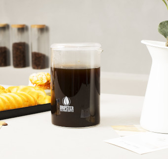 Mit Kaffee gefülltes Dripster Gefäß, im Hintergrund Gebäck und drei Gläser mit Kaffeebohnen