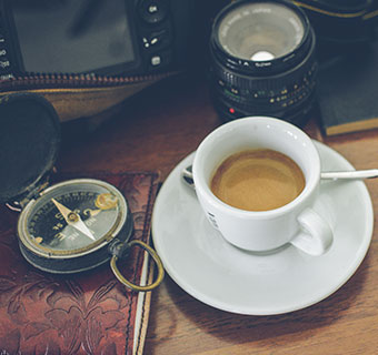 Weiße Espressotasse steht auf Holztisch neben einem Kompass und einem Kameraobjektiv