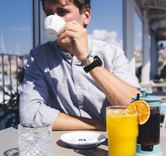 Mann sitzt am Café-Tisch in der Sonne und trinkt aus weißer Cappuccinotasse mit blauem Bazzara Schriftzug